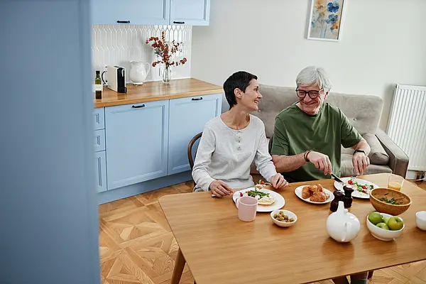Älteres Paar isst lächelnd Frühstück an einem Holztisch in einer hellen Küche.