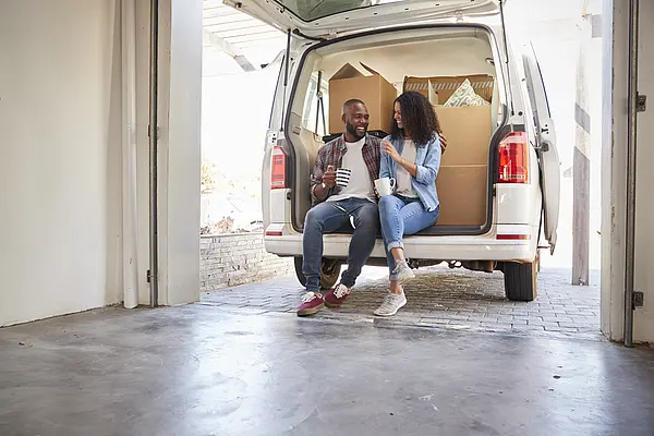 Paar sitzt entspannt auf Lieferwagen, umringt von Umzugskartons, im offenen Garagentor.