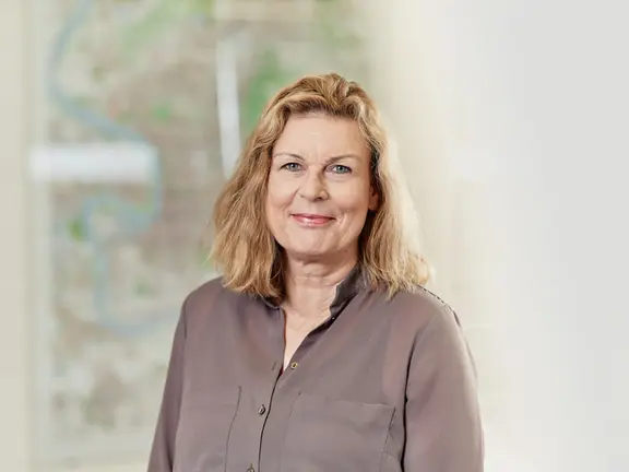 Frau mit blonder Frisur, lächelt, vor unscharfem Hintergrund mit Karte.