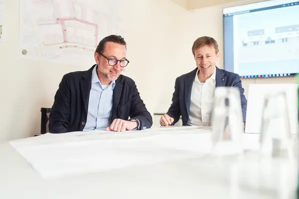 Zwei Männer sitzen an einem Tisch mit Plänen und Gläsern, im Hintergrund ein Bildschirm.