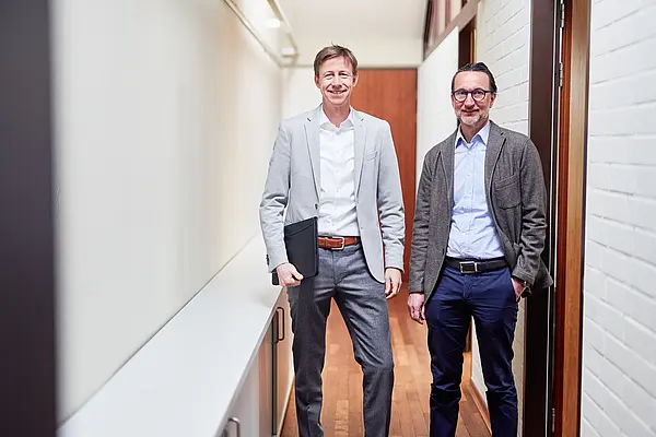 Zwei Männer in einem Flur, elegant gekleidet und lächelnd, einer hält einen Laptop.