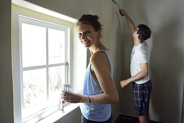 Zwei Personen streichen eine weiße Wand neben einem sonnigen Fenster.