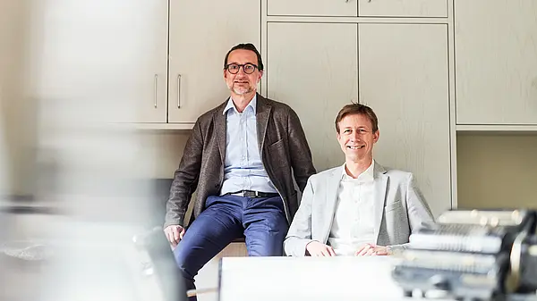 Zwei Männer in einem Büro, einer sitzt am Tisch, der andere lehnt an einem Schrank.