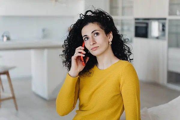 Frau mit lockigem Haar telefoniert nachdenklich im Wohnzimmer.