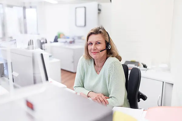 Frau mit Headset sitzt im Büro und lächelt in die Kamera.